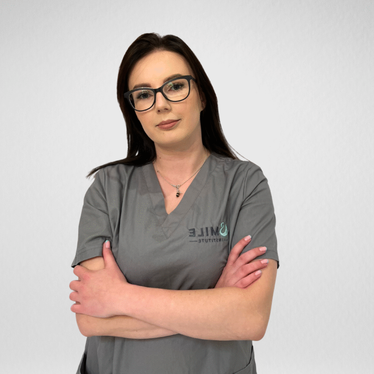 Higienistka Stomatologiczna Karolina Pszczoła z rękami skrzyżowanymi na klatce piersiowej w szarym fartuchu z logo Smile Institute